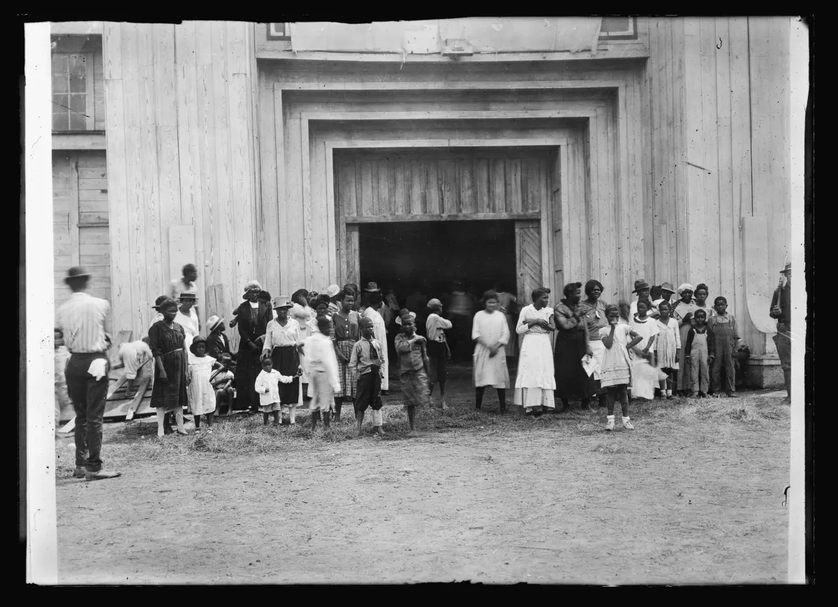 Black-and-white photograph of the entrance to the Tulsa fairgrounds refugee camp, June 1921. A wooden gate is flanked by makeshift signage. Black Tulsans displaced by the massacre stand near the entrance; tents and camp structures are visible in the background.