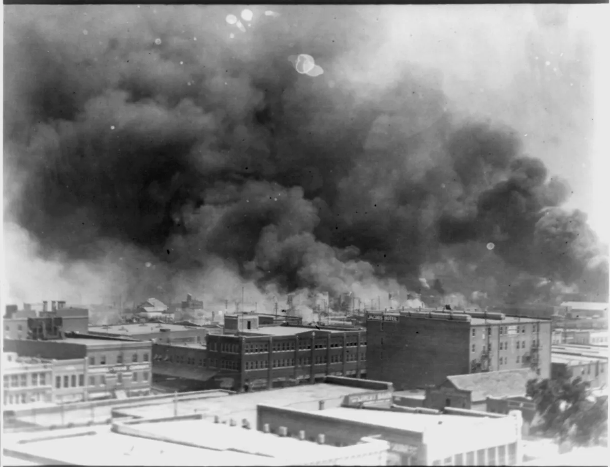 Black-and-white aerial photograph of dense smoke rising over Tulsa rooftops on the morning of June 1, 1921. Multiple plumes of black smoke billow above a dense city skyline; flames are visible at street level in the foreground.