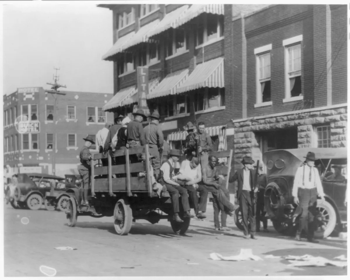 Black-and-white photograph of a flatbed truck on a Tulsa street near the Litan Hotel. Armed white men stand on the truck bed alongside Black Tulsans who have been detained. A handful of figures stand in the road around the truck.