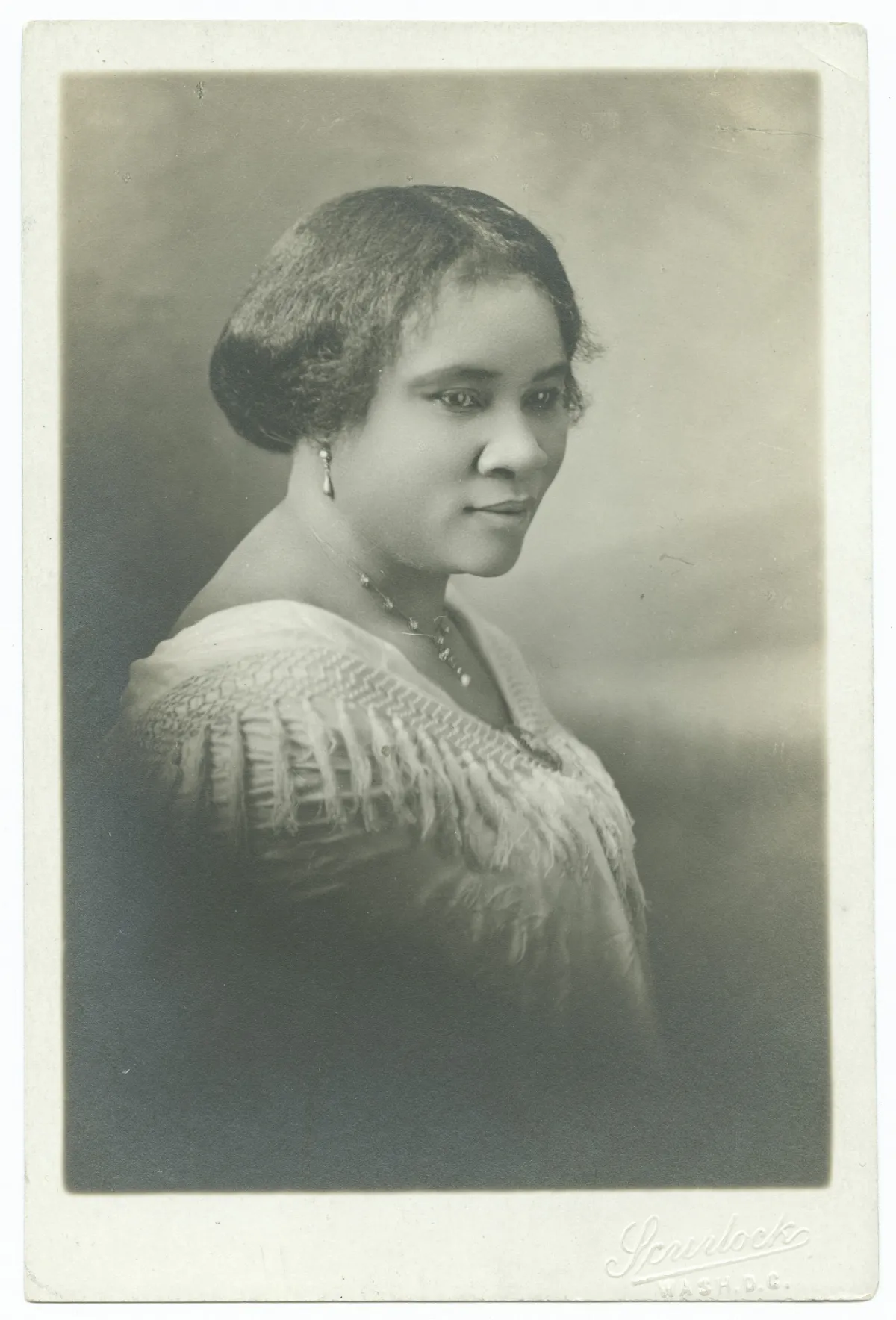 Black-and-white studio portrait of Madam C.J. Walker, c. 1915. Walker is seated, looking directly at the camera, in a tailored dark dress with high lace collar. The lighting is soft and frontal in the Scurlock studio style.